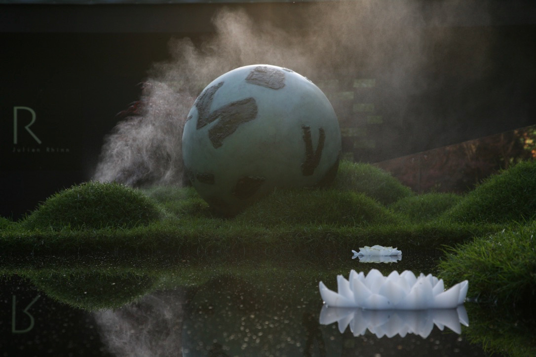 Bronze sphere sculpture in situ on moss with mist and water reflection